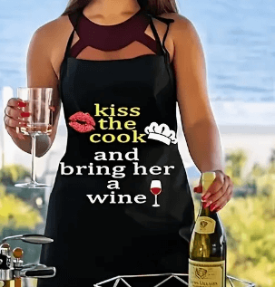 Apron Kiss The Cook Wine Apron worn by a person holding a glass and a bottle of wine outdoors.